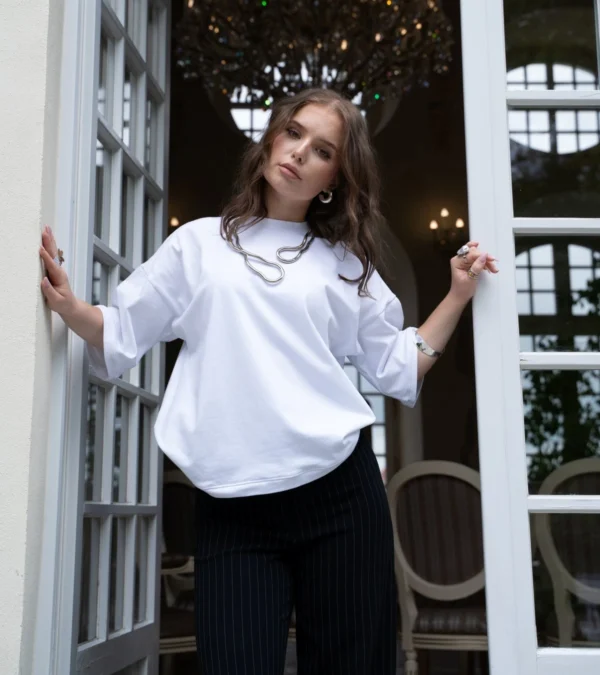 Biała koszulka oversize z bawełny o szerokich rękawach. Modelka prezentuje ją w eleganckiej stylizacji z czarnymi spodniami w prążki.